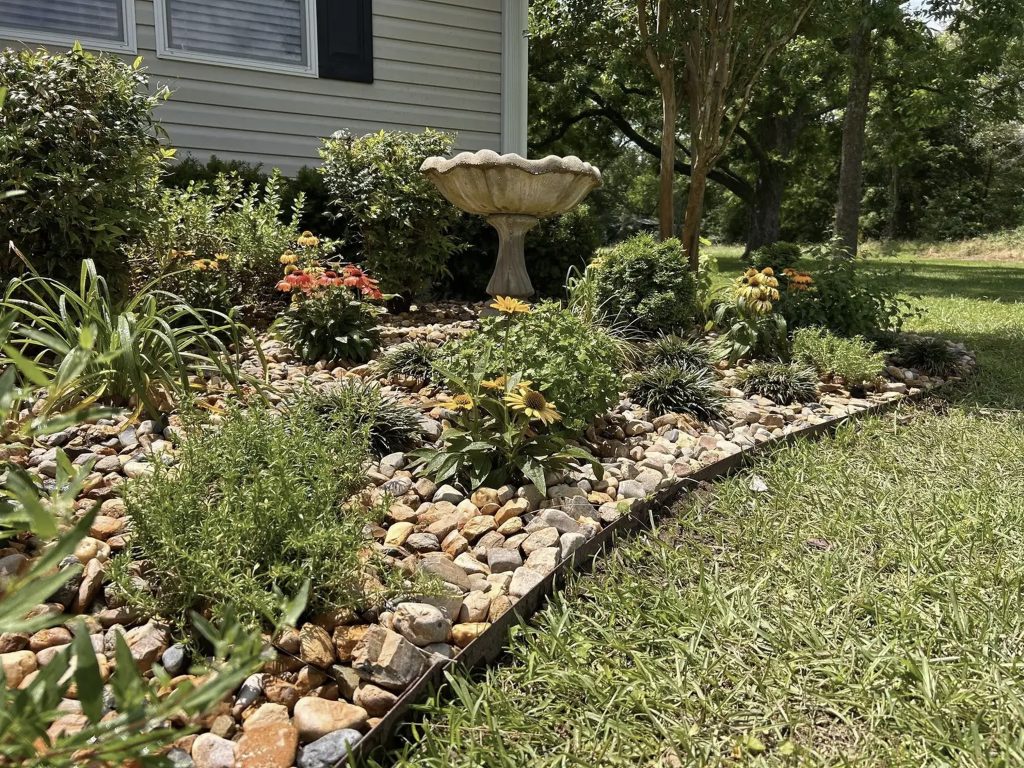 Front yard landscaping with bird bath, decorative rock garden, and blooming flowers by Hood Lawn Care
